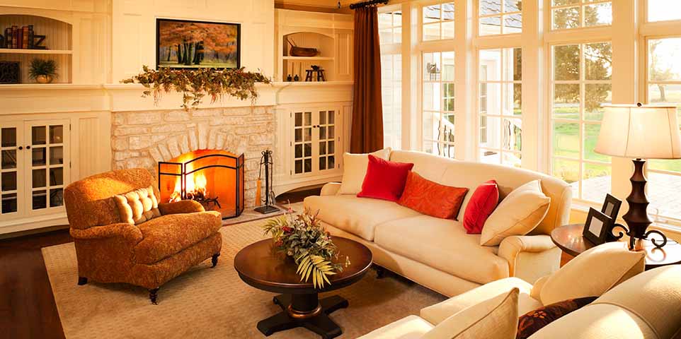 Cozy living room with a lit fireplace, beige sofa with red cushions, brown armchair, glass coffee table with a flower arrangement, large windows letting in natural light, and warm-toned decor.