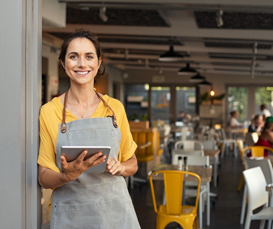 Alt tag: Small business owner wearing apron and holding a tablet inside a modern café or restaurant. Alt tag: Small business owner wearing apron and holding a tablet inside a modern café or restaurant.