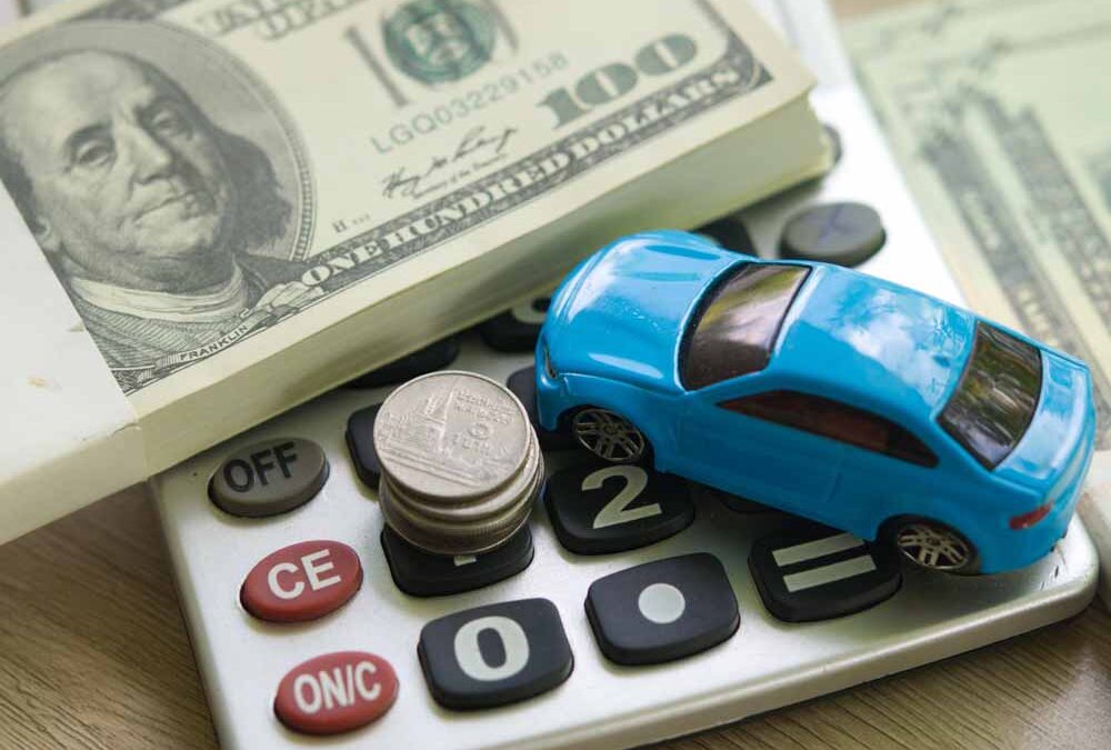 Blue toy car on a calculator with stacked coins and US hundred-dollar bills in the background.