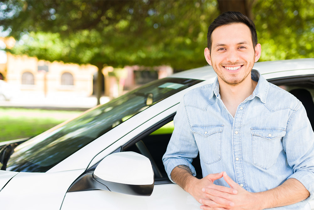 A smiling man in a light blue denim shirt leans against a white car in a sunny outdoor setting with blurred green trees in the background