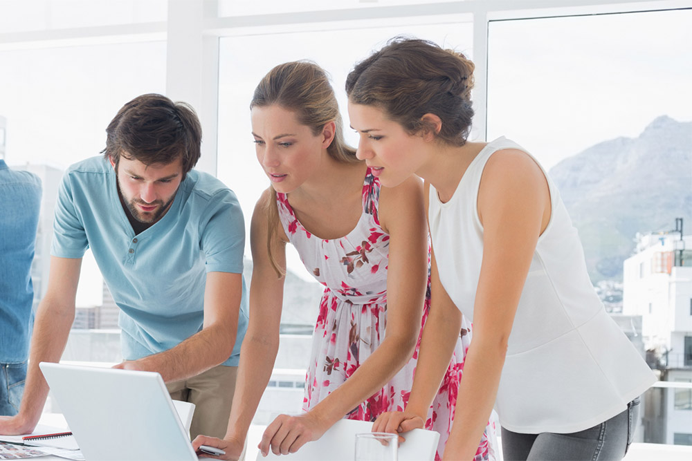 Three colleagues leaning over a desk to collaborate and look intently at a laptop screen in a bright, modern office
