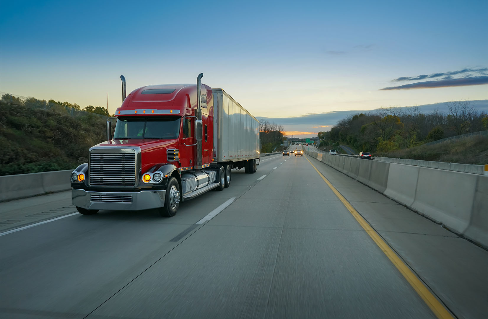 A red semi-truck speeding down a highway between concrete barriers during early morning or evening twilight