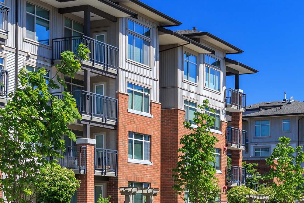 A modern multi-story apartment building with red brick and grey siding features black metal balconies and large windows under a clear blue sky