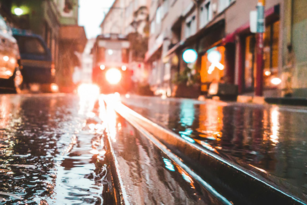 A low-angle shot of a wet city street, with tram tracks reflecting the bright headlights of an approaching, blurred red tram and glowing storefronts