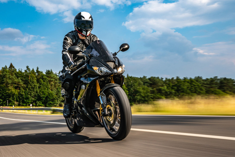 A motorcyclist wearing black leathers and a helmet rides a black sportbike at high speed down a sunny highway