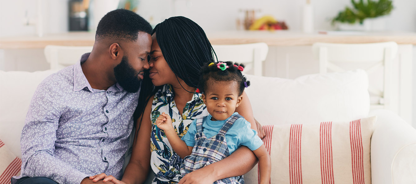 A Black father and mother touch foreheads affectionately while their young daughter sits on her mother's lap and smiles at the camera