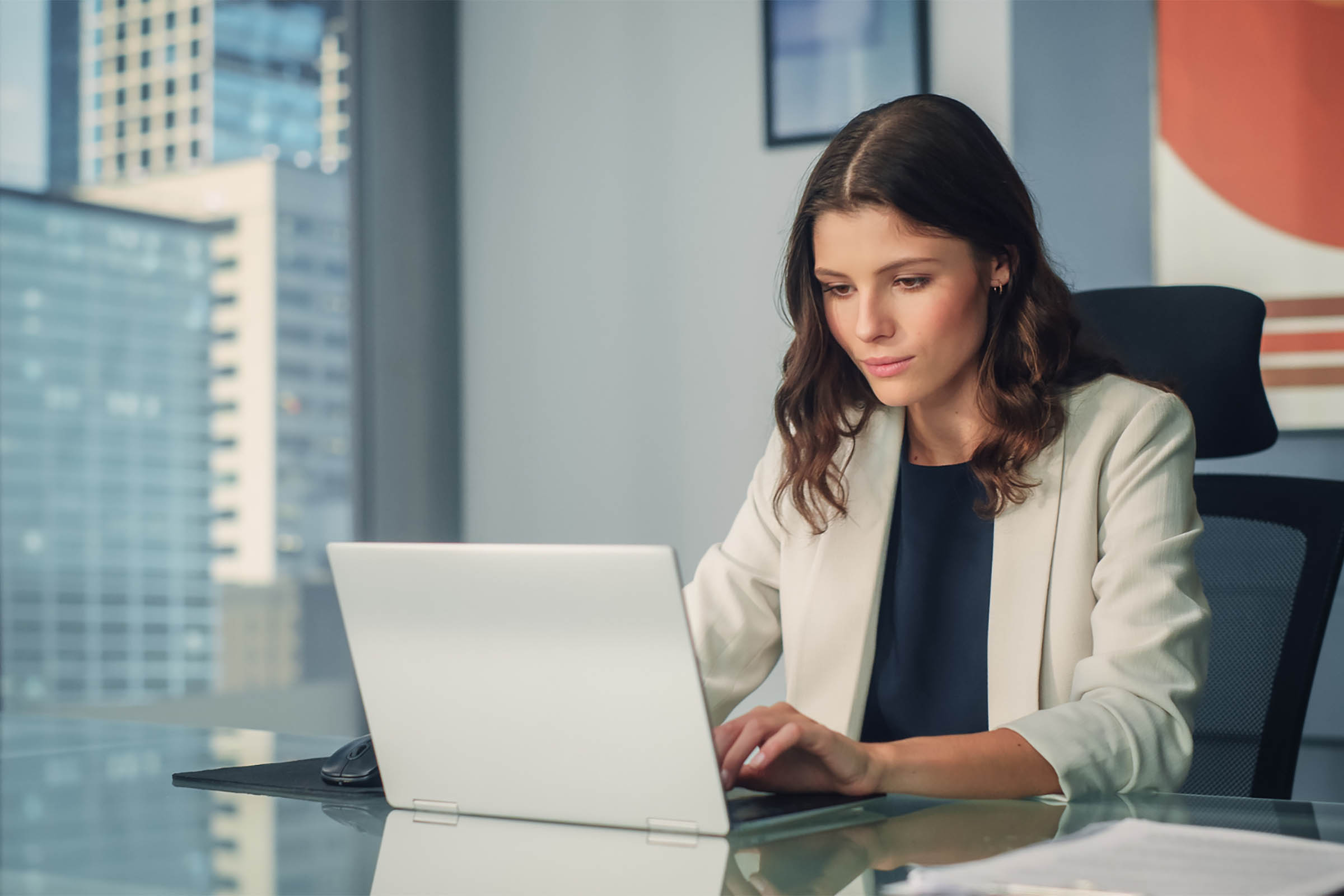 A focused professional woman in a white blazer working on a silver laptop at a glass desk in a modern corporate office