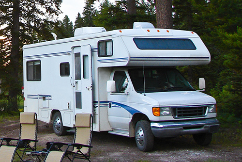 A white motorhome with blue stripes parked in a wooded campsite, with its side door open and two camping chairs set up outside