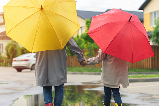 A mother and daughter, walking away from the camera on a wet street after rain, each holding a vibrant umbrella—one yellow and one red