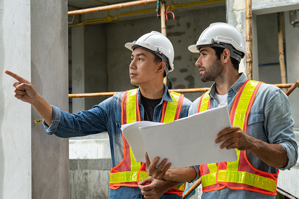 Two construction workers in hard hats stand in front of a partially constructed building, discussing their project